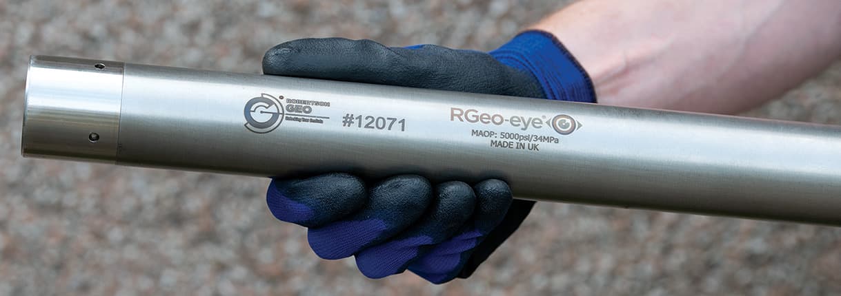 Metal downhole camera probe held by a gloved operator, with RGeo-eye markings. Reference equipment photograph.