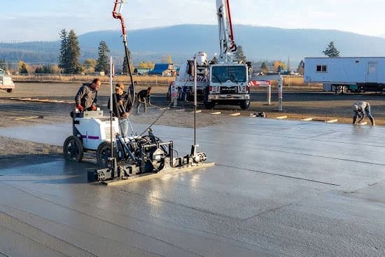 Construction-site photograph of operators guiding a compact laser screed with two upright receiver masts across wet concrete; concrete placing booms appear behind. Reference equipment photograph.