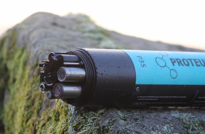 Black Proteus multiparameter probe with water droplets resting on a rock; several exposed sensing elements are visible. Reference equipment photograph.