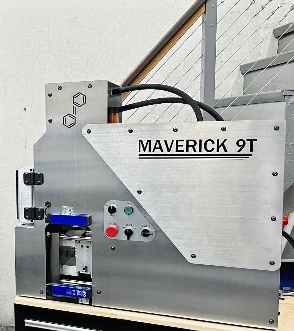 Polymer Dynamics Maverick 9T compact injection molder showing the enclosed body, mold opening and control buttons. Reference equipment photograph.